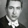 Black and white image of a young man wearing a black robe or jacket with a white bow tie. He looks at the camera