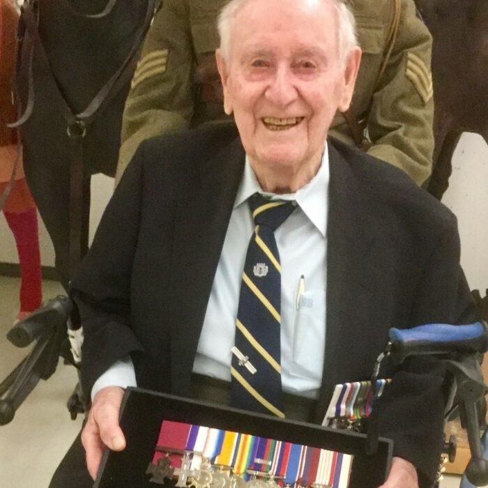 William Little with His Medals, Date Unknown, The Fort Garry Horse Museum and Archives A colour image of an old man seated holding a case full of war medals. He is wearing a suit and tie over a white button up shirt. He smiles at the camera
