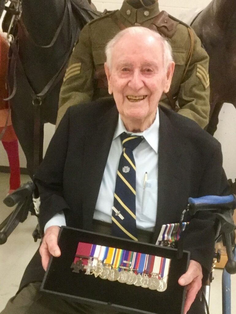 A colour image of an old man seated holding a case full of war medals. He is wearing a suit and tie over a white button up shirt. He smiles at the camera