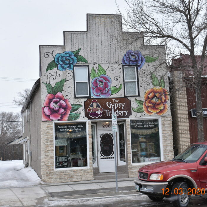 A colour photo of a white stucco building. The facade of the building is made in the 'boom-town' style and there are murals of roses painted above the door and around the windows. There are two upper-story windows and one large bay window split by the front door on the main level