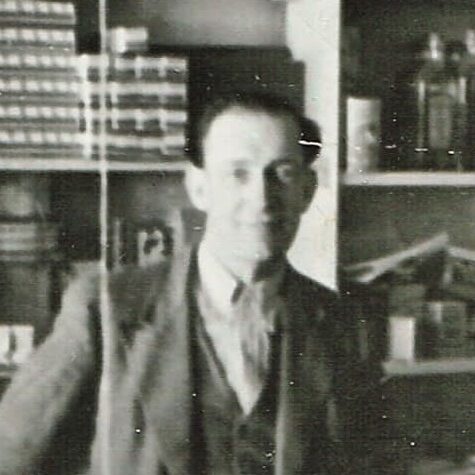 Black and white photo of a man standing at a shop counter. In the background are goods for sale
