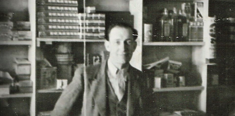 Black and white photo of a man standing at a shop counter. In the background are goods for sale