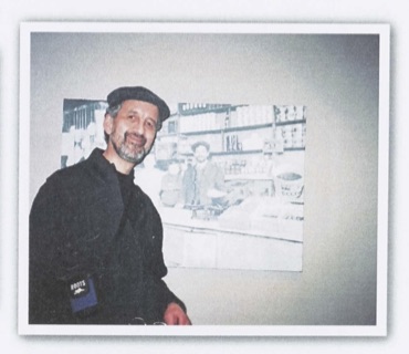 Colour photo of a man standing next to a photo of a young child. The man wears a jacket and a hat. He has a beard