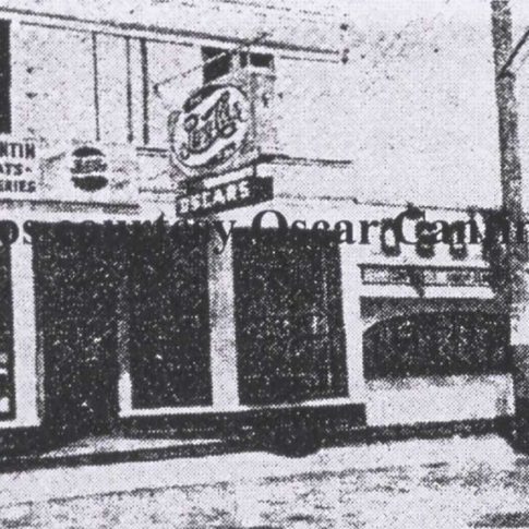Black and white photo of a two-story building. The main floor has windows and the second floor has a hanging sign