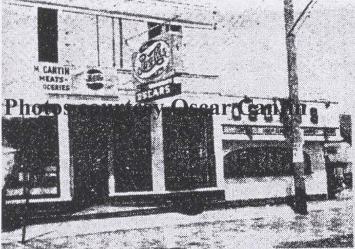 Black and white photo of a two-story building. The main floor has windows and the second floor has a hanging sign