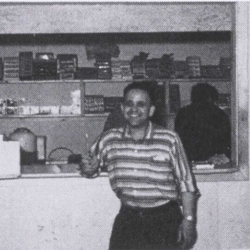 Black and white photo of a man standing in front of a shop counter. He is wearing a striped shirt and black pants. In the background are shelves with boxes and various items on it