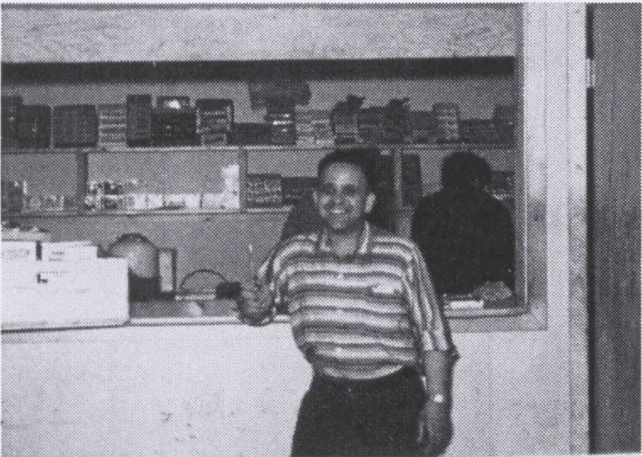 Black and white photo of a man standing in front of a shop counter. He is wearing a striped shirt and black pants. In the background are shelves with boxes and various items on it