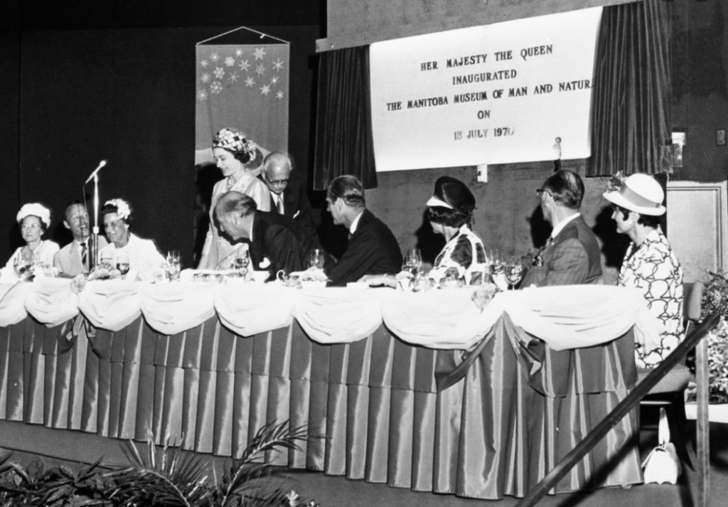 A black and white photo of a row of people sitting at a long decorated table. The table has a shiny, dark tablecloth and a white topper tablecloth. The people are seated and facing one woman who is standing on the left. In the background there is a large white sign hanging as well as a banner