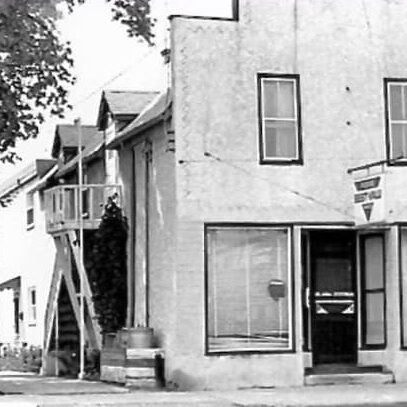 A black and white photo of a white stucco building. The facade of the building is made in the 'boom-town' style. There are two windows on the second floor and large bay windows on either side of the door on the first floor
