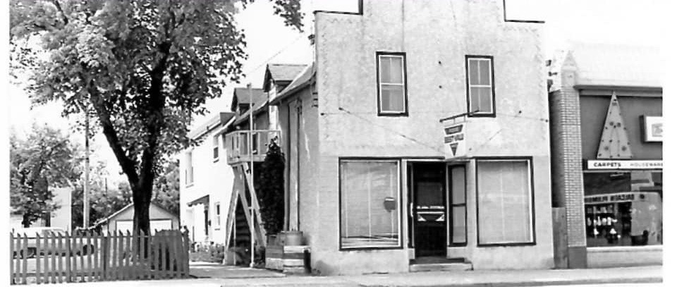A black and white photo of a white stucco building. The facade of the building is made in the 'boom-town' style. There are two windows on the second floor and large bay windows on either side of the door on the first floor