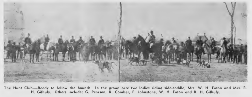 Black and white photo of a row of men sitting on horseback. In the foreground there are two dogs running across the frame