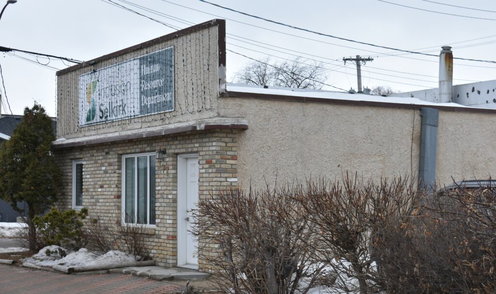 A colour photo of a beige building. The photo is taken from a three quarter perspective. There are leafless shrubs along the side of the building as it is winter-time. The building has christmas lights on the top, front portion of the boom town facade style building.