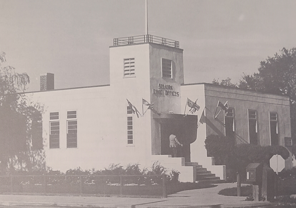 Black and white photo of Civic Office. The building is white and has a flag pole on the top. There are smaller flags on the wall around the entrance to the building