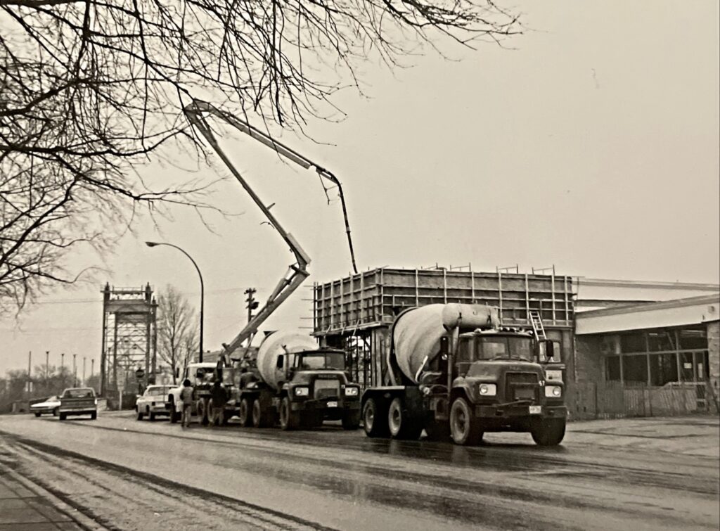 Black and white photo of the construction of Civic Office. There is a large pump truck parked outside the building and a cement truck driving away down the road