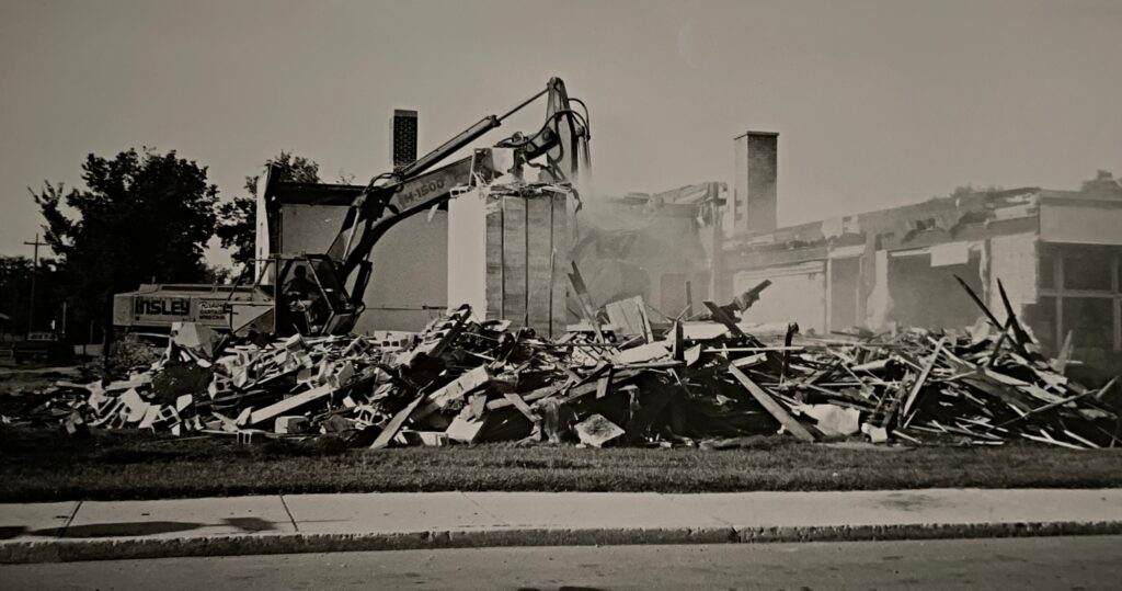 Black and white photo of the demolition of the old Civic Office. There is an excavator on the left side of the photo and lots of rubble and dust at the center of the frame.