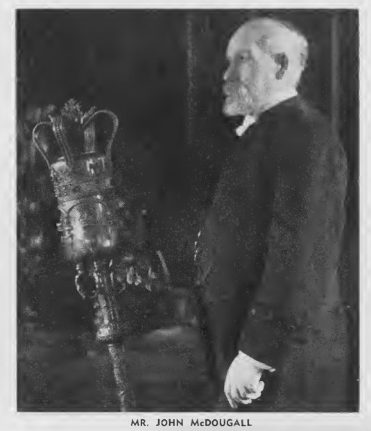 Black and white photo of a man wearing a suit. Hes standing next to a large crown on a scepter
