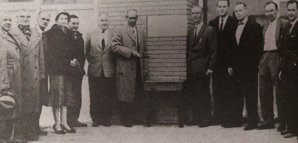 Black and white photo of a a group of men standing around a brick wall and plaque