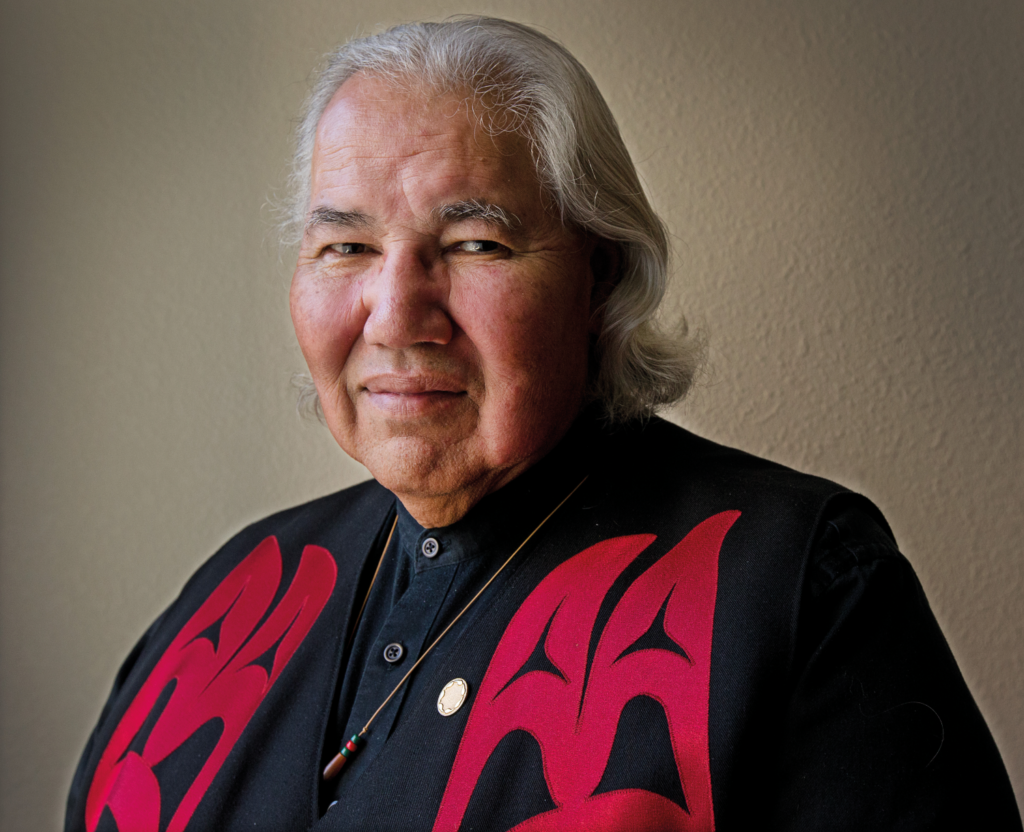 Colour photo of a man seated looking at the camera. He has shoulder-length grey hair and is wearing a black shirt with red Indigenous patterning on the chest