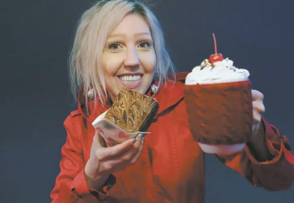 Colour photo of a woman holding some baking and a drink with whipped cream and a cherry on top. She has blonde hair and is smiling. She is also wearing a bright red jacket