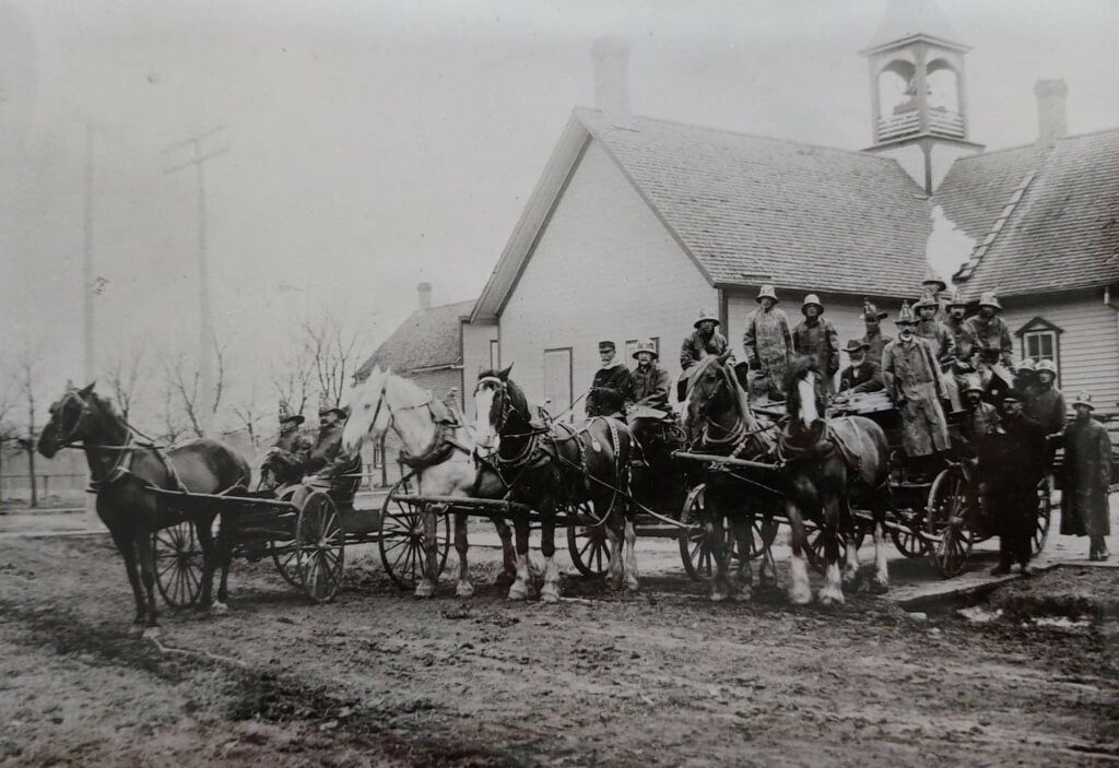 Black and white photo of town hall and the fire department in the front of the building. There are horses, wagons, and firefighters standing on the wagons