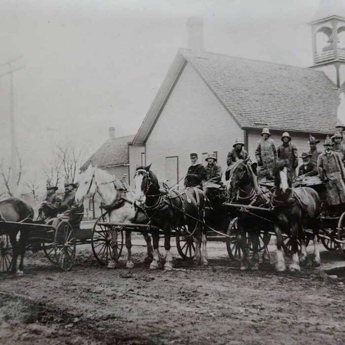 The First Fire Wagons with View of Selkirk Town Hall in the Background, 1906, Ted Wozny Black and white photo of town hall and the fire department in the front of the building. There are horses, wagons, and firefighters standing on the wagons