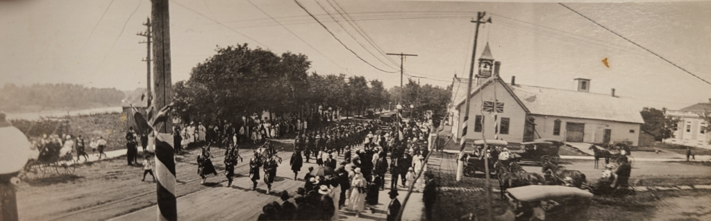 Black and white photo of a parade making its way down the street. Town Hall is visible on the right side of the photo