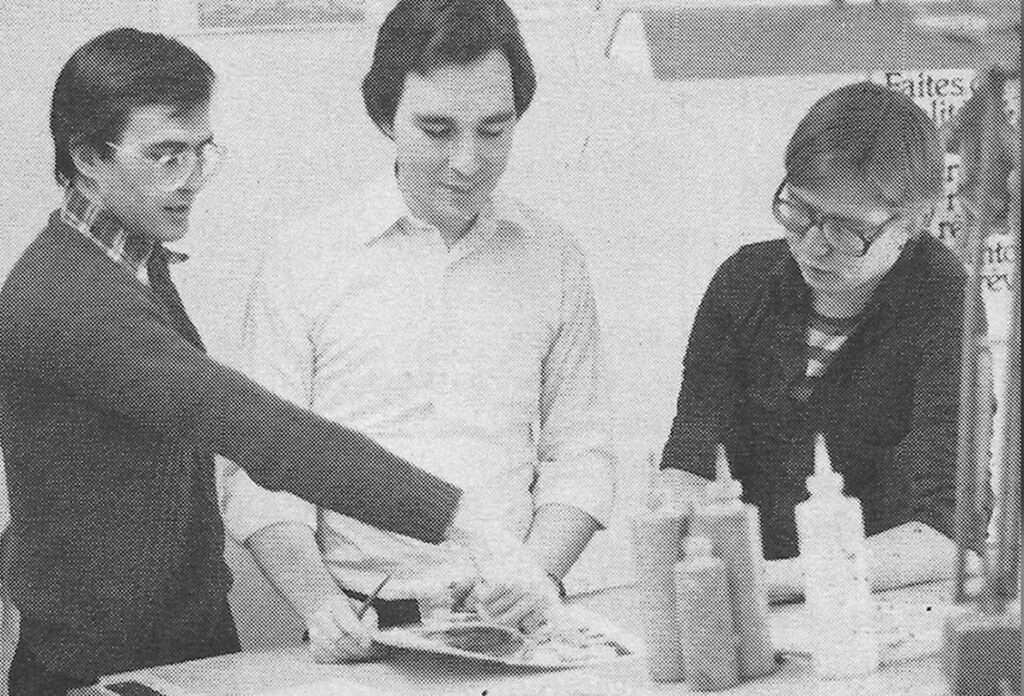 Black and white photo of three men standing together working at a table