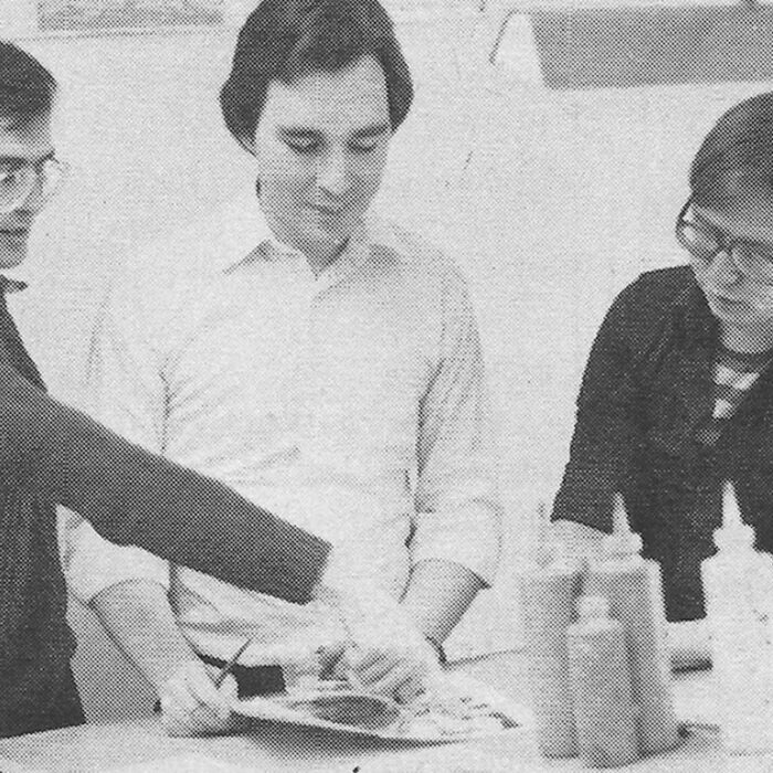 Black and white photo of three men standing together working at a table