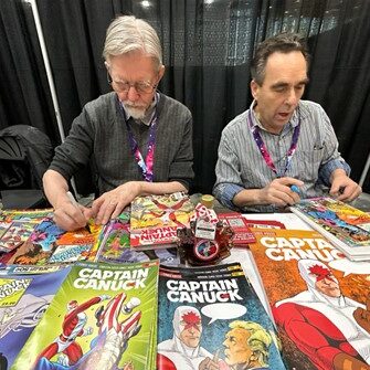 Colour photo of two men sitting at table at a comiccon booth signing comic books