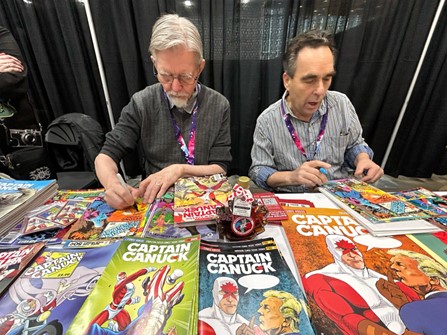 Colour photo of two men sitting at table at a comiccon booth signing comic books
