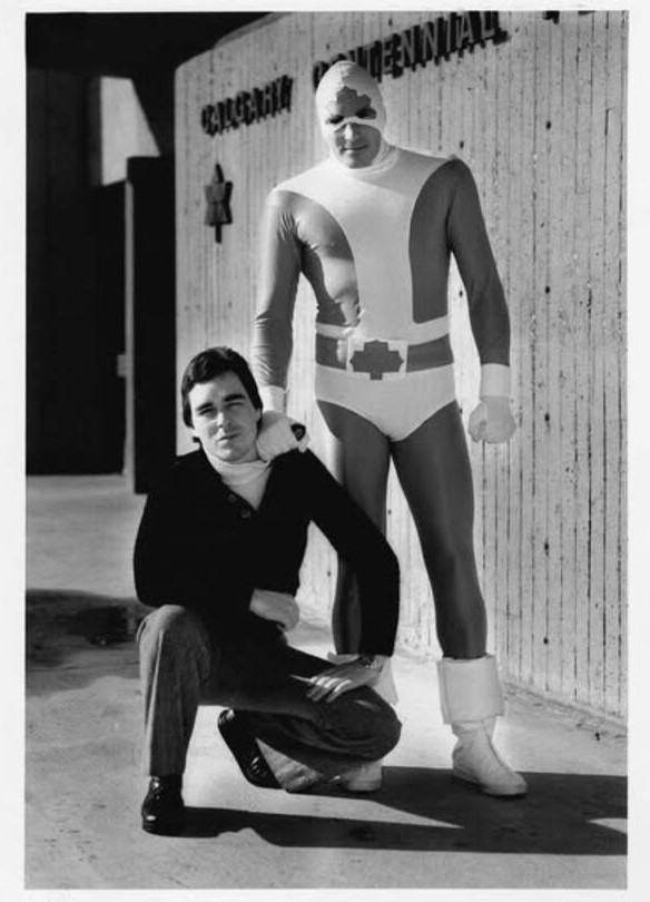Black and white photo of Richard Comely crouching next to a man dressed as Captain Canuck