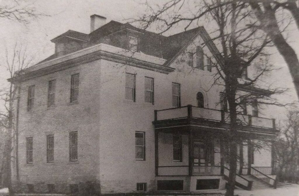 Black and white photo of a three story building. It is white with a dark peaked roof