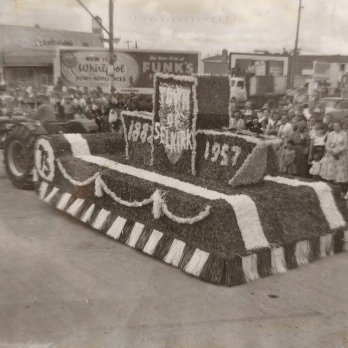 Selkirks 75th Parade Town of Selkirk, 1957, Selkirk Museum Collection Black and white photo of the Town of Selkirk's parade float. The floar is decorated with flowers and a long banner which runs the length of the flat deck trailer