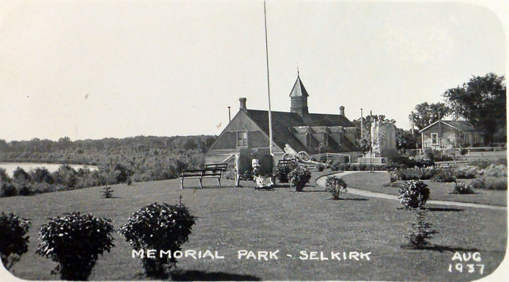 Black and white photo of a building on the far side of a grassy park. There are shrubs in the foreground and a flag pole at the center of the image