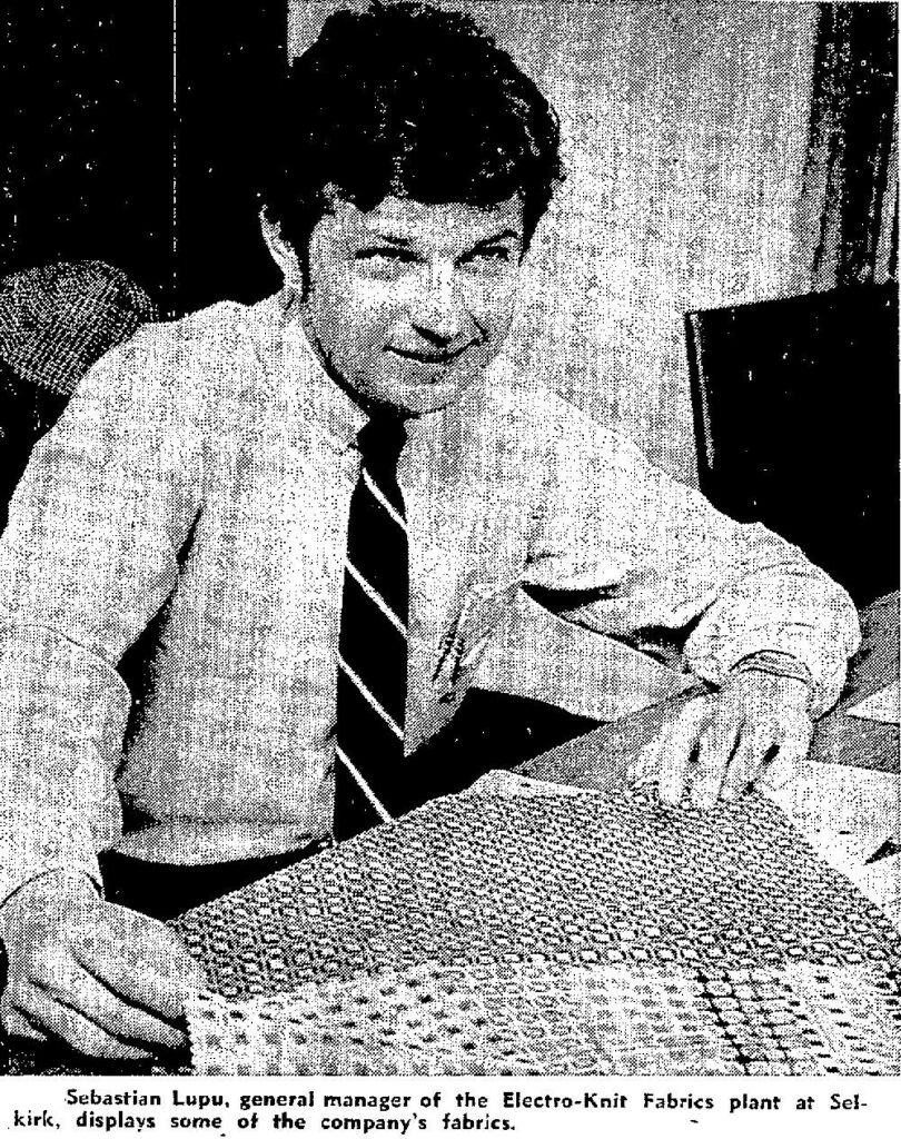 Black and white photo of a man seated at a desk . He is wearing a white shirt with a striped tie. He looks to the right and smiles