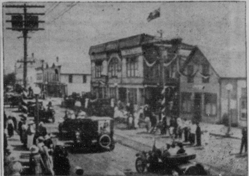 Black and white photo of a two story building with banners strung on the outside and a flag flying on the top of the building